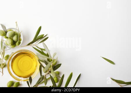 Bol en verre rempli d'huile d'olive et de branches d'olive isolées sur une table blanche. Vue de dessus. Banque D'Images