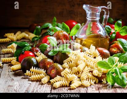 Pâtes italiennes sèches spirales, basilic vert, huile d'olive dans une carafe, tomates cerises noires et rouges, encore la vie dans un style rustique sur fond de bois vintage Banque D'Images