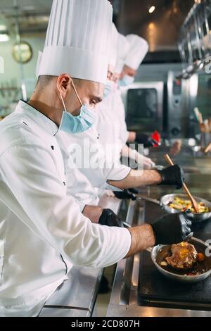 Les chefs cuisiniers en masques et gants de protection préparent des aliments dans la cuisine d'un restaurant ou d'un hôtel. Banque D'Images