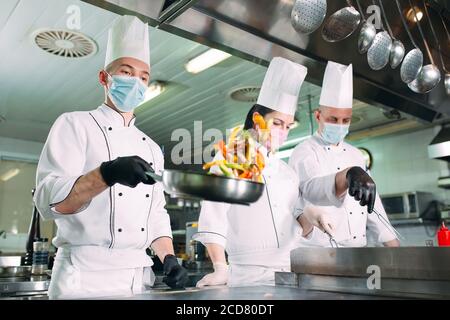 Les chefs cuisiniers en masques et gants de protection préparent des aliments dans la cuisine d'un restaurant ou d'un hôtel. Banque D'Images