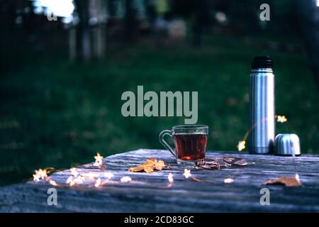 Pique-nique d'automne dans le parc avec une tasse de thé chaud, des thermos, des lumières de noël et des feuilles jaunes sur une table rustique en bois à l'extérieur . Banque D'Images