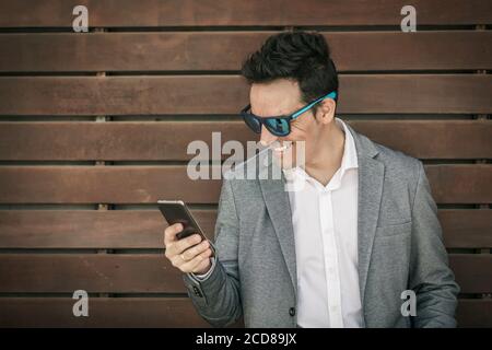 Happy élégant homme en costume élégant et lunettes de soleil tendance debout contre un mur en bois et la lecture d'actualités sur internet via smartphone Banque D'Images