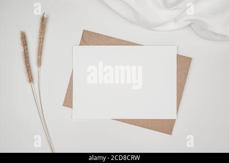 Papier blanc vierge sur une enveloppe en papier marron avec une fleur sèche et un chiffon blanc. Maquette de carte de vœux vierge horizontale. Vue de dessus du CRAF Banque D'Images