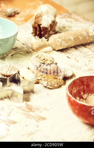 délicieux cookies au chocolat aux copeaux saupoudrés de sucre en poudre à proximité de bols turquoise rouges, d'un couteau et d'une broche à rouler sur une table en bois avec beaucoup de farine, foyer sélectif Banque D'Images