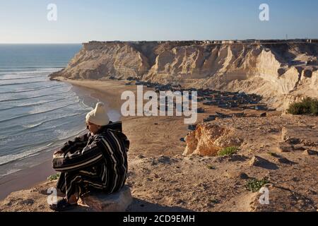 Maroc, Sahara occidental, Dakhla, vieil homme assis sur une falaise surplombant la plage d'Araiche et ses bateaux de pêche Banque D'Images