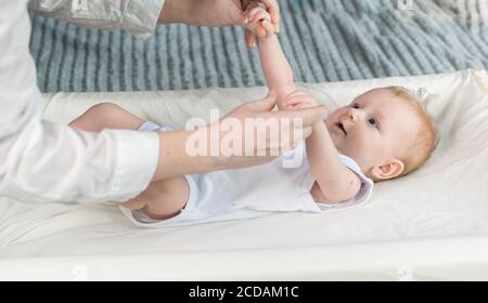 Les mains des infirmières font de la gymnastique pour un bébé magnifique dans un body blanc. Le bébé est allongé sur la table à langer Banque D'Images