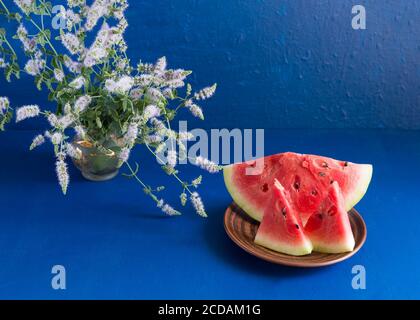 Trois tranches de pastèque fraîche mûre sont dans une assiette en argile sur fond bleu foncé. Dans un vase en verre, un bouquet d'origan. Copier l'espace Banque D'Images