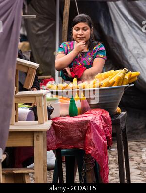 Une jeune femme maya Quiche en robe traditionnelle vend de l'elote ou du maïs cuit à la vapeur sur l'épi dans le marché de Chichichastenango, au Guatemala. Banque D'Images