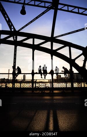 Personnes mal identifiées et rétroéclairées appréciant le coucher du soleil assis sur un pont en acier sur des voies de chemin de fer appelées Hackerbrücke. Munich, Allemagne. Banque D'Images