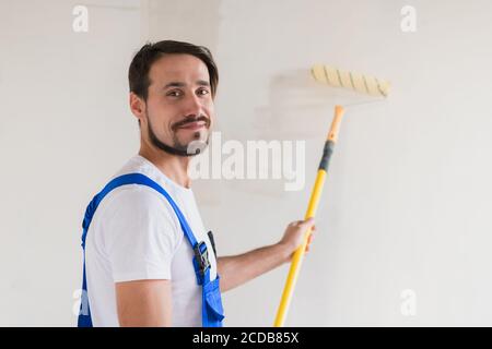 Un homme barbu en vêtements de travail peint le mur avec un rouleau. Il sourit amical et pose pour la caméra Banque D'Images