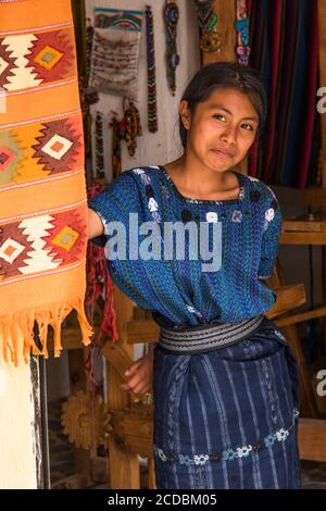 Une jolie jeune fille Maya portant robe typique regarde par la porte de l'atelier de tissage de la famille à San Antonio Palopo, au Guatemala. Banque D'Images