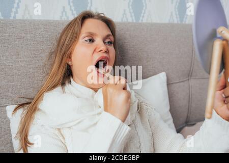 Jolie blonde femme vérifiant sa gorge à l'aide de bâton de bois et miroir couché dans le lit et se sentant malade. Banque D'Images