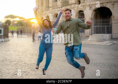 Jeunes couples de touristes prenant des photos sautant près du colisée avec minuterie à rome au coucher du soleil. Couple romantique de jeunes touristes à l'écoute de la musique et Banque D'Images