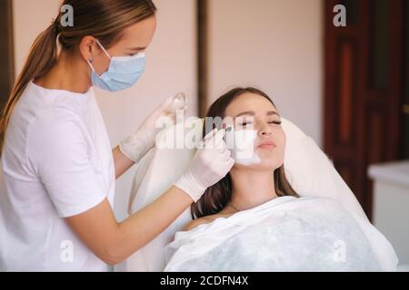 Jeune femme cosmetologiste appliquer le masque blanc d'argile sur le visage de la femme. Belle femme brune dans le salon de beauté Banque D'Images