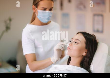 Jeune femme cosmetologiste appliquer le masque blanc d'argile sur le visage de la femme. Belle femme brune dans le salon de beauté Banque D'Images