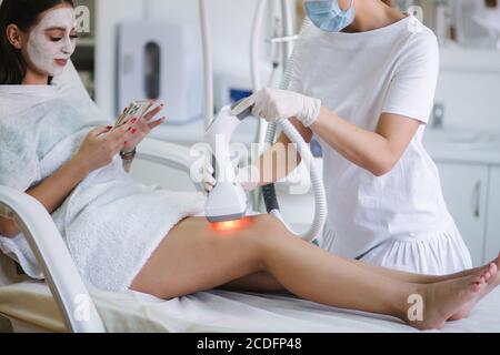 Une femme de bussines beutiful avec un masque facial blanc utilisant le téléphone pendant cosmétologue en gants blancs effectuant une procédure de levage par radiofréquence sur le Banque D'Images