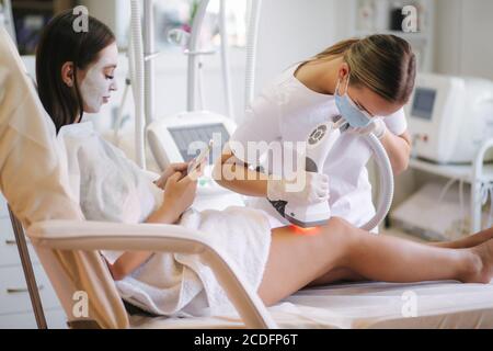 Une femme de bussines beutiful avec un masque facial blanc utilisant le téléphone pendant cosmétologue en gants blancs effectuant une procédure de levage par radiofréquence sur le Banque D'Images