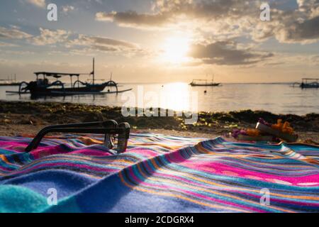 Lever du soleil sur Sanur Beach Bali avec des lunettes et du canang en premier plan. Un bateau traditionnel appelé un jukung se prépare à mettre la voile. Une matinée incroyable Banque D'Images