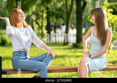 Deux jeunes filles amis assis sur un banc dans le parc d'été discutant heureusement avoir du plaisir.