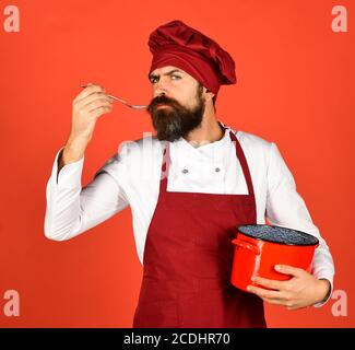 Un homme avec une barbe tient les ustensiles de cuisine sur fond rouge. Concept du processus de cuisson. Chef avec une casserole ou une casserole rouge et une cuillère. Faites cuire avec un visage sérieux dans la soupe au chapeau bordeaux et au tablier Banque D'Images