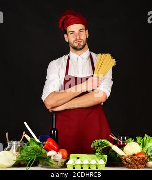 Ustensiles de cuisine et concept de cuisine. Le chef au visage plein de confiance tient un tas de spaghetti sur fond noir. L'homme en chapeau et en tablier tient les pâtes. Cuisiner travaille dans la cuisine près de la table avec des légumes et des outils. Banque D'Images