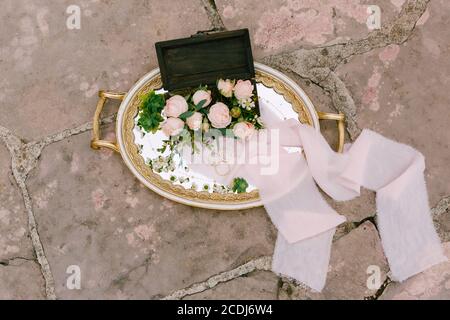 Anneaux de mariage et un anneau d'engagement sur un plateau avec des motifs et un bouquet de roses roses roses dans une boîte en bois. Banque D'Images