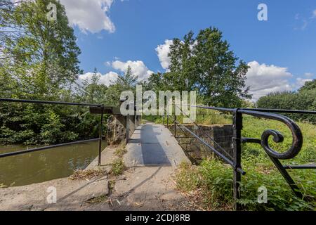 Petit pont avec sa balustrade en métal noir au-dessus du barrage et de la lisière du ruisseau Keutelbeek entouré de végétation verte dans le parc de la ville de Sittard, ensoleillé Banque D'Images