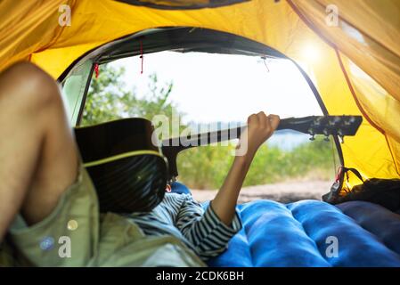 Adolescent allongé dans une tente jouant de la guitare. Jeune homme qui apprécie les loisirs en plein air près de la rivière. Camping. Vacances d'été. Concept de tourisme local Banque D'Images