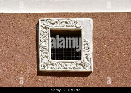 Petite ventilation carrée avec grille de poulet et cadre de fenêtre en plâtre blanc orné. Isolé sur un mur en pointillé de galets de couleur sable. Banque D'Images