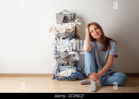 Jeune femme aux cheveux rouges assise sur le parquet avec une pile de garde-robe et du linge dans un panier à linge en métal. Banque D'Images