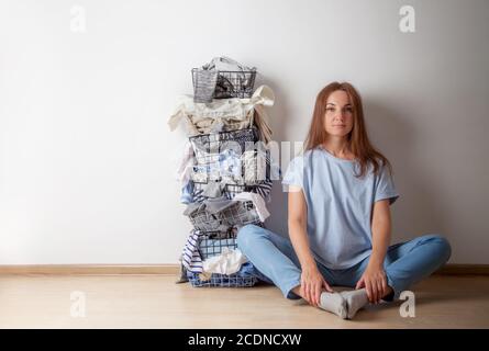 Jeune femme aux cheveux rouges assise sur le parquet avec une pile de garde-robe et du linge dans un panier à linge en métal. Banque D'Images