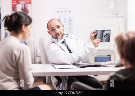 Médecin montrant une blessure au genou sur une image radiographique dans le bureau de l'hôpital à une jeune femme et à sa mère handicapée en fauteuil roulant. Medic portant une couche blanche et un stéthoscope. Banque D'Images