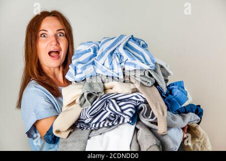 Surprise jeune femme aux cheveux rouges portant une pile de vêtements encombrés à la maison. Banque D'Images