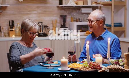 Surprise femme âgée avec un sourire charmant assis à la table dans la cuisine ouvrant petite boîte cadeau présent. Joyeux couple dînant ensemble à la maison, appréciant le repas, célébrant leur anniversaire Banque D'Images