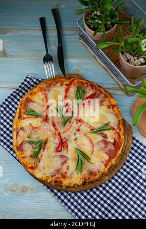 Pizza hawaïenne traditionnelle sur un plateau en bois sur la table avec une serviette à carreaux. Pizza ronde avec viande, poivre, saucisse, romarin et fromage Banque D'Images