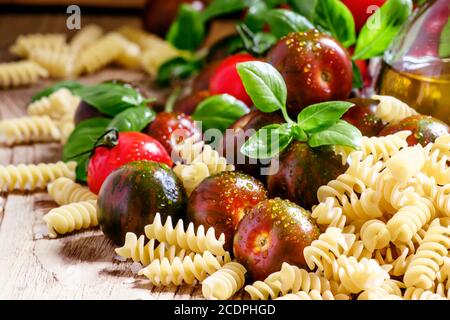 Pâtes italiennes sèches spirales, basilic vert, huile d'olive dans une carafe, tomates cerises noires et rouges, encore la vie dans un style rustique sur fond de bois vintage Banque D'Images