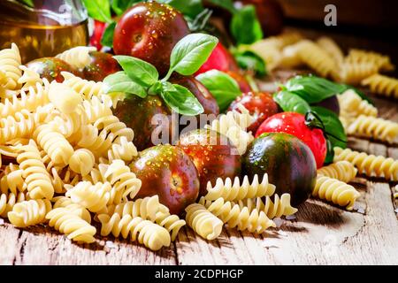 Pâtes italiennes sèches spirales, basilic vert, huile d'olive dans une carafe, tomates cerises noires et rouges, encore la vie dans un style rustique sur fond de bois vintage Banque D'Images