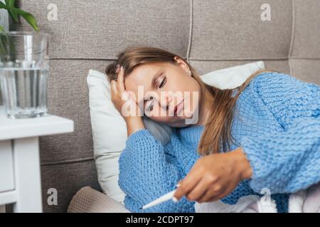 Une femme malade se trouve dans un lit sous une couverture et vérifie le thermomètre. Maux de tête, grippe, rhume, virus. Banque D'Images