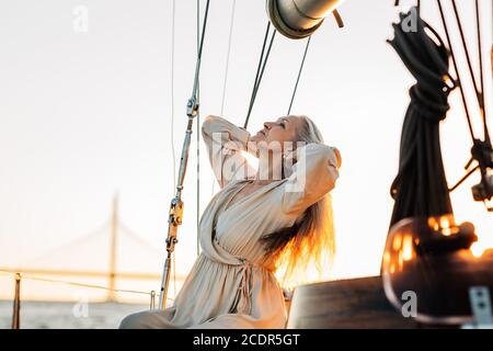 Vue latérale d'une femme âgée élégante assise sur un yacht terrasse et ajuster ses cheveux longs au coucher du soleil Banque D'Images