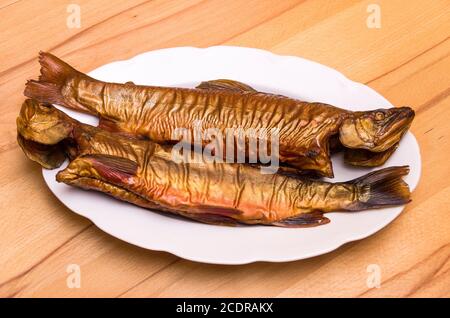 Deux poissons fumés, l'omble de fontaine, sur une assiette blanche sur une table en hêtre. Banque D'Images