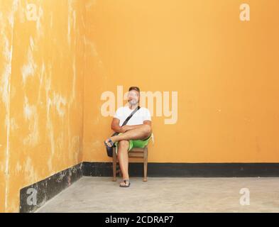 Jeune homme assis sur une chaise en bois dans un délabeur chambre avec murs orange Banque D'Images