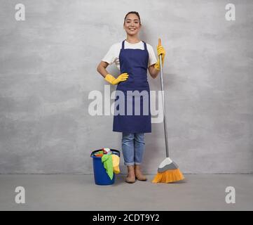 Meilleurs services de nettoyage. Bonne jeune femme de nettoyage en uniforme et gants en caoutchouc tenant le balai et le seau avec différents produits de nettoyage, regardant l'appareil photo et souriant, debout contre le mur gris Banque D'Images