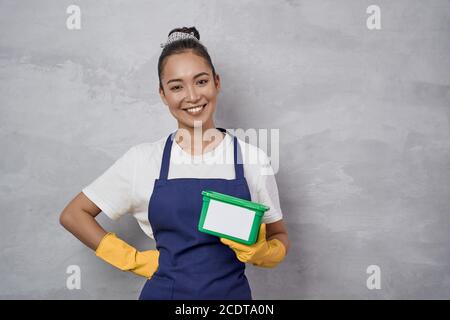 Le concept de ménage et de ménage. Portrait d'une jeune femme de ménage souriante ou femme de ménage en uniforme et gants en caoutchouc jaune tenant une boîte en plastique vert avec des capsules de lavage, regardant l'appareil photo Banque D'Images