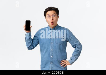 Surpris et stupéfait asiatique homme dans des lunettes et des vêtements décontractés, montrant l'écran de téléphone mobile et de dire wow, discutant de nouvelle application de smartphone, enfin Banque D'Images