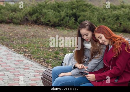 Les adolescentes d'écouter de la musique sur smartphone assis sur le banc dans un parc de la ville d'automne Banque D'Images