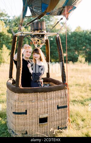 Beau jeune couple souriant en amour, portant des vêtements décontractés noirs, rester et embrasser dans le panier ballon d'air au coucher du soleil, prêt pour leur premier Banque D'Images