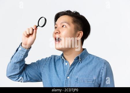 Gros plan d'un homme asiatique fasciné, chercheur en chemise bleue, regardant à travers la loupe dans le coin supérieur gauche avec une expression stupéfaite, trouvé Banque D'Images