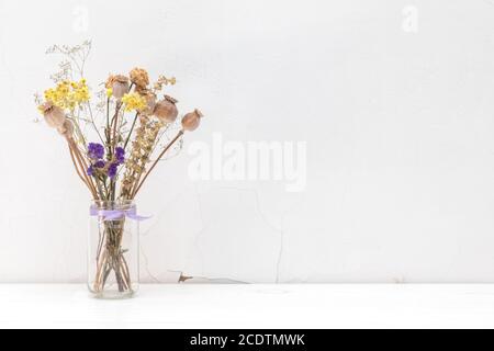 Fleurs séchées et têtes de pavot dans un pot en verre sur fond de mur blanc fissuré. Banque D'Images