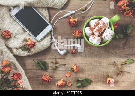 Tasse de café avec guimauve, laine à tricoter blanche, fleurs de roses séchées, téléphone portable et casque Banque D'Images