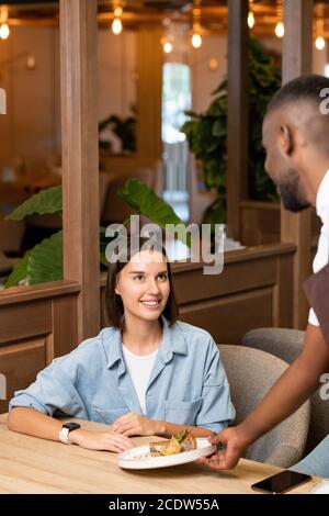 Jeune femme souriante en chemise bleue regardant le serveur africain service à elle Banque D'Images